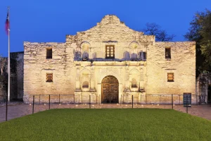 Illuminated exterior of the Alamo at dusk with a flag visible to the side