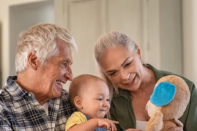 Grandparents playing with a laughing baby holding a teddy bear at home