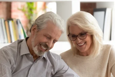 Older man signing a document while his smiling wife looks on at a table