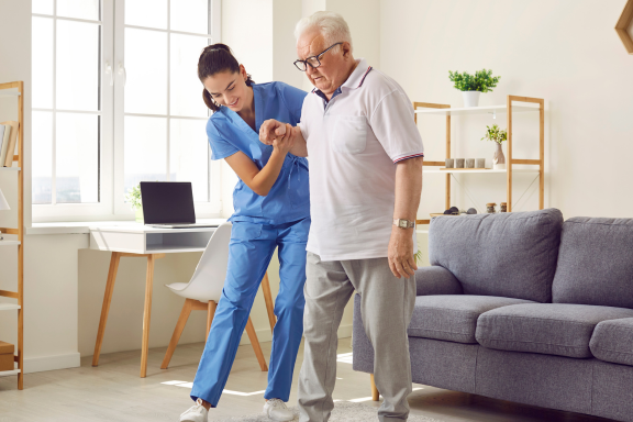 Caregiver in scrubs helping an older man walk across a living room