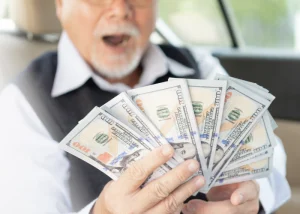 Man in a car holding a fanned stack of hundred-dollar bills, looking excited
