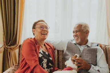 Smiling couple seated on a striped sofa, gesturing toward their small dog