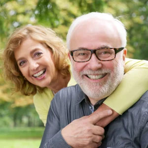 Older man laughing outdoors while a woman peeks over his shoulder smiling