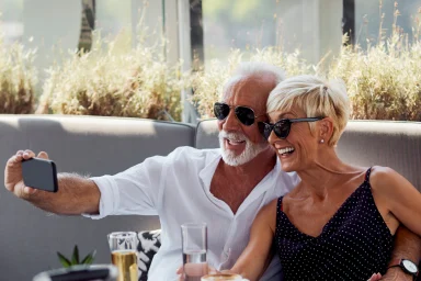 Older couple taking a selfie at an outdoor cafe table with a suitcase nearby