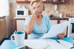 Older woman at a kitchen table covering her face while holding paperwork