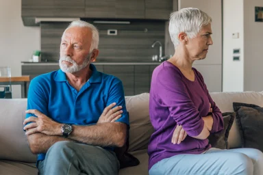 Older couple sitting apart on a couch with arms crossed, looking away from each other