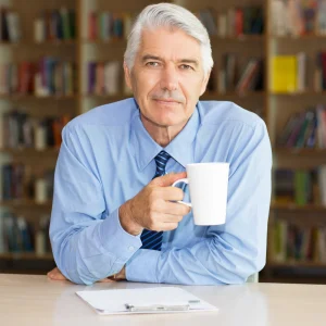 Older man in a home library holding a coffee mug with a clipboard nearby