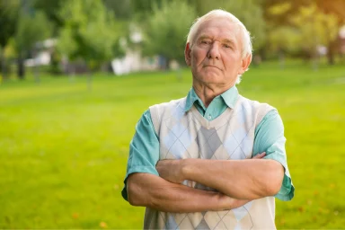 Older man with arms crossed standing in a sunny park, looking stern
