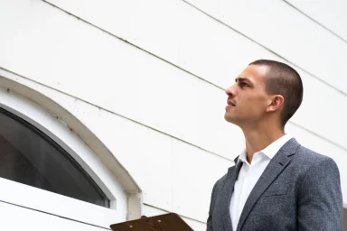 Home inspector in a suit holding a clipboard while examining a building's siding