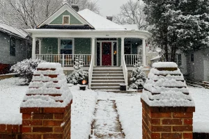 Snow-covered green house with a lit porch and brick gateposts in winter