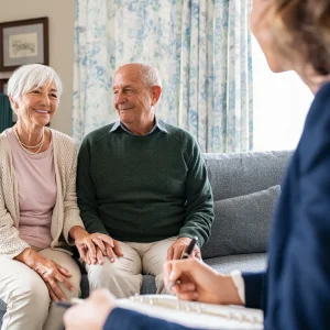 Older couple smiling and holding hands on a sofa while an advisor takes notes