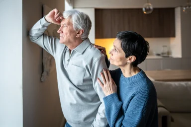 Older man looking out a window while his wife rests a hand on his shoulder