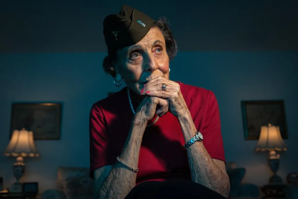 Elderly woman in a military garrison cap sitting with hands clasped