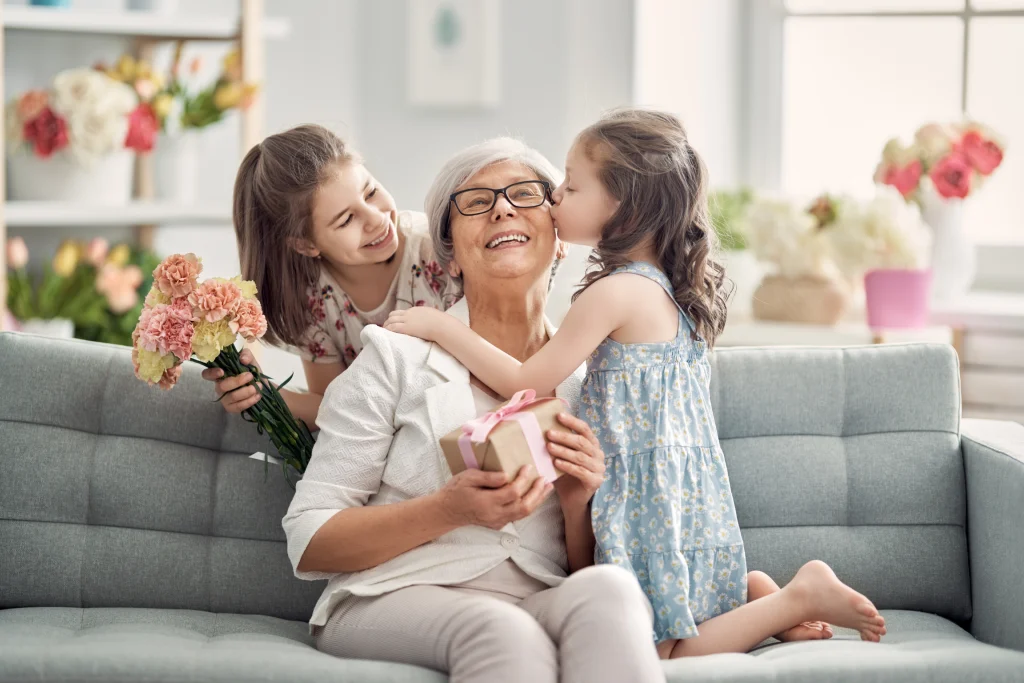 Grandmother on a couch receiving flowers and a kiss from her granddaughters