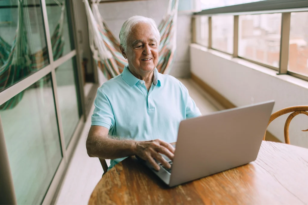 Smiling older man typing on a laptop at a table on a sunlit balcony