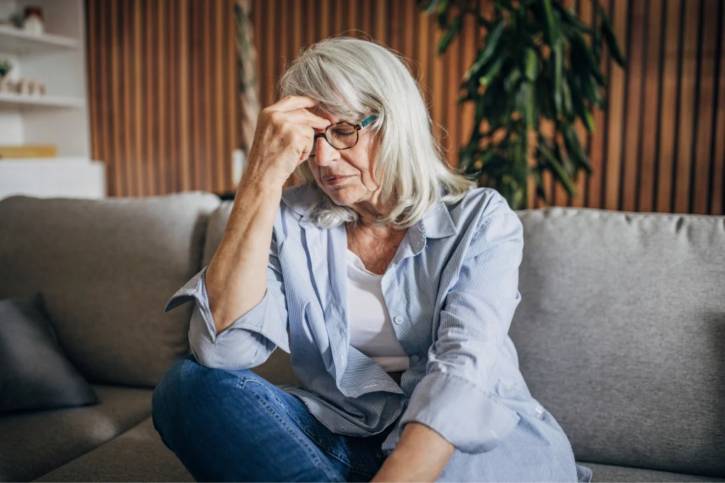 Older woman sitting on a couch looking stressed with her hand to her forehead