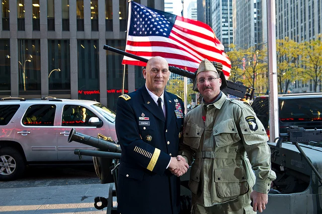 Uniformed serviceman shaking hands with a WWII-era reenactor by a flag