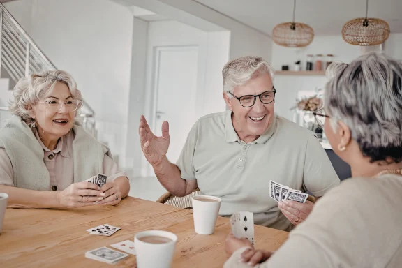 Three older adults laughing while playing cards at a wooden table