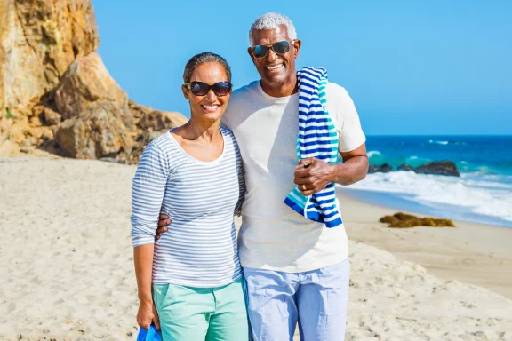 Older couple smiling with arms around each other on a sandy beach