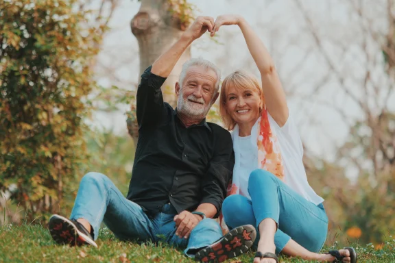 Older couple sitting on grass forming a heart shape with their raised hands